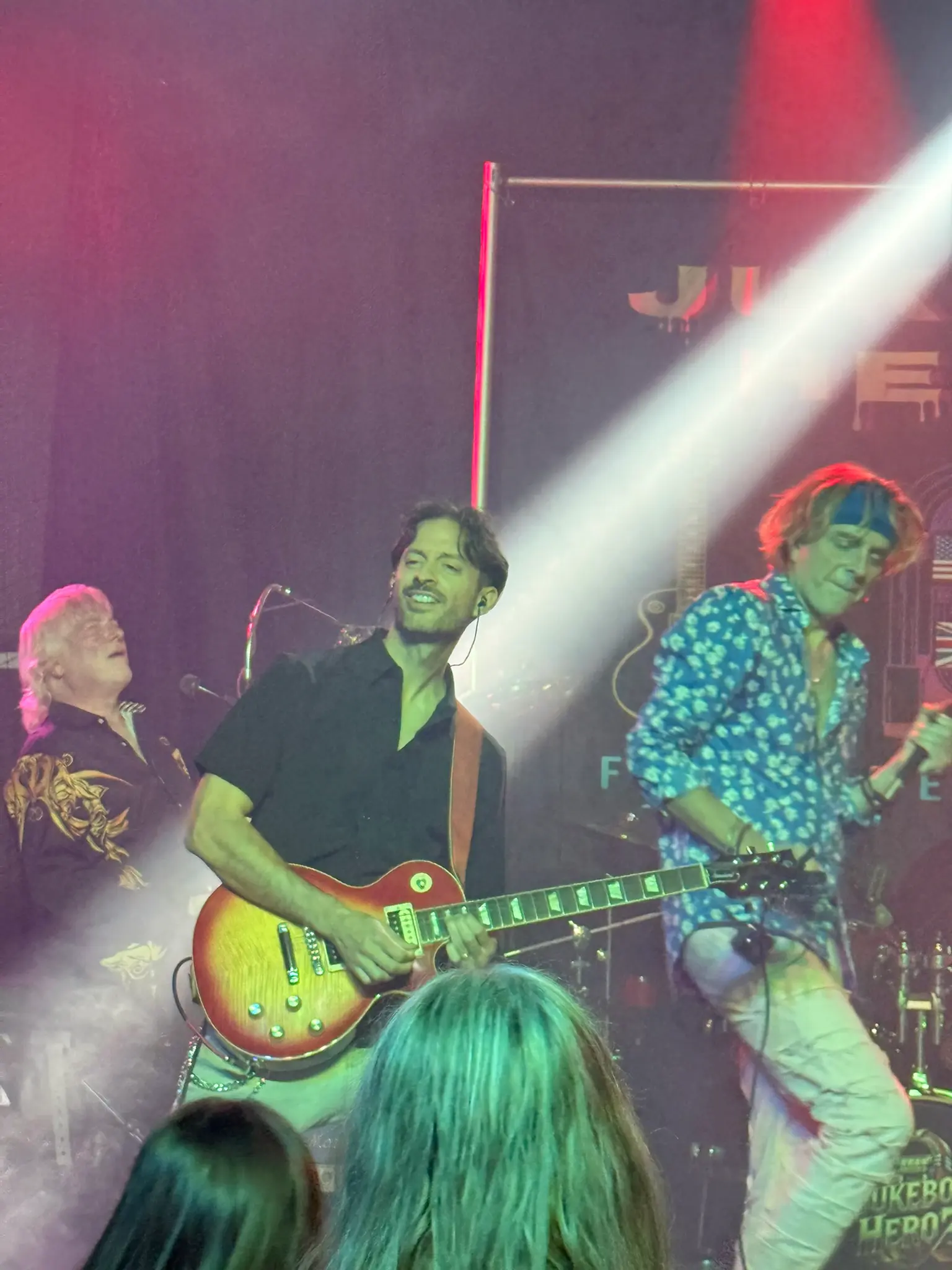 Jukebox Hero'z guitarist playing a sunburst Les Paul beside the singer, caught in a beam of white stage light