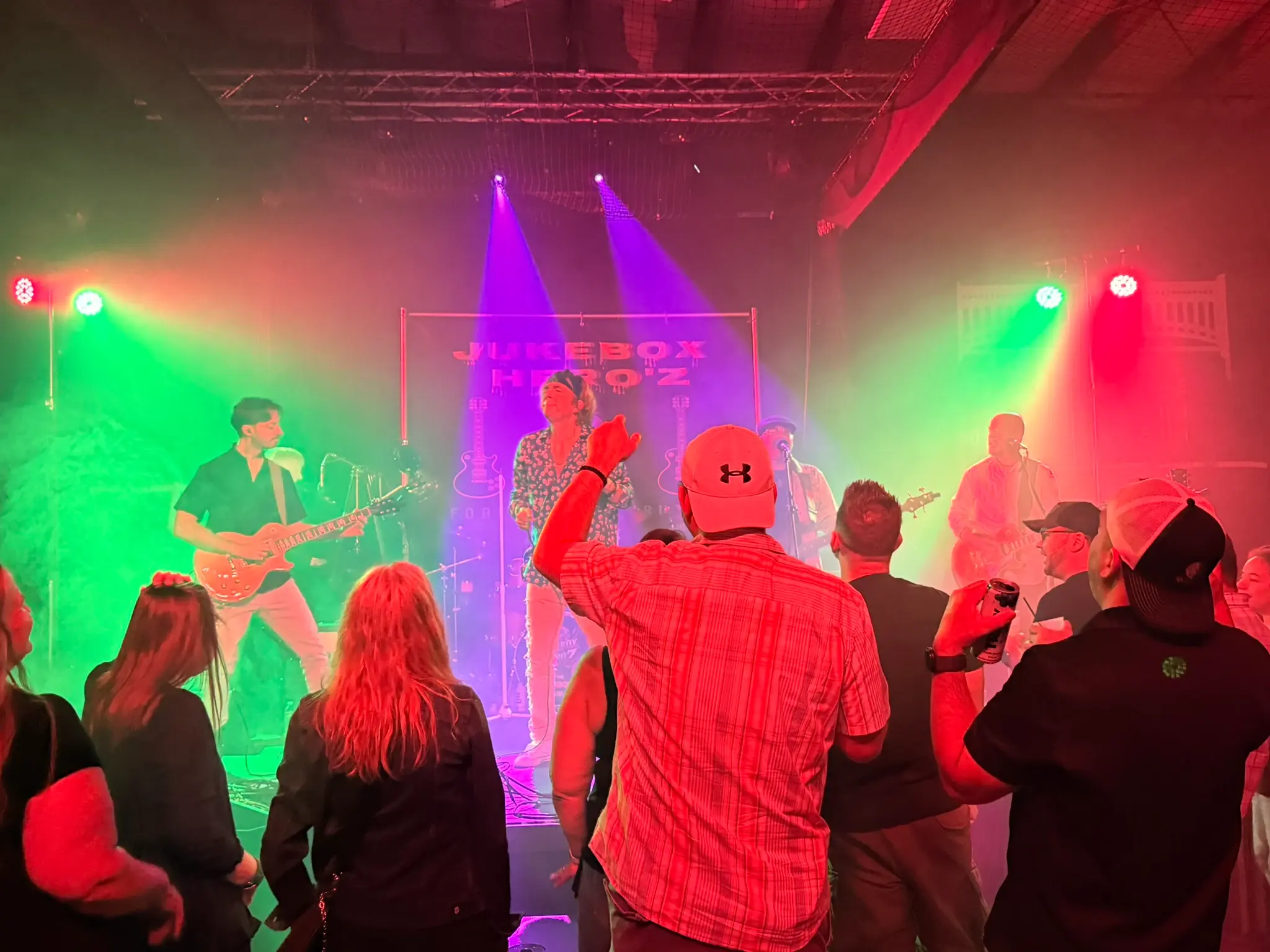 A packed crowd watching Jukebox Hero'z perform under green and red stage lights, the band's banner behind the stage